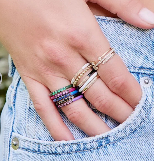 A hand with multiple rings rests casually in a blue denim pocket. Among them is the Rachel Reid Black Enamel And Pave Diamond Band Ring, complemented by colorful stacked bands, all set in 14K yellow gold—highlighting a playful and stylish accessory choice.