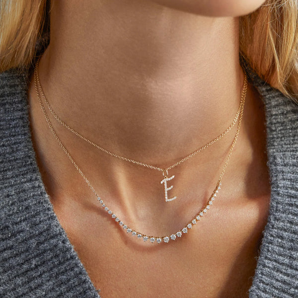 A woman in a gray knit top wears two delicate 14k yellow gold necklaces by ef collection: one featuring the Diamond Script Initial Charm and another adorned with small, evenly spaced diamond gemstones.