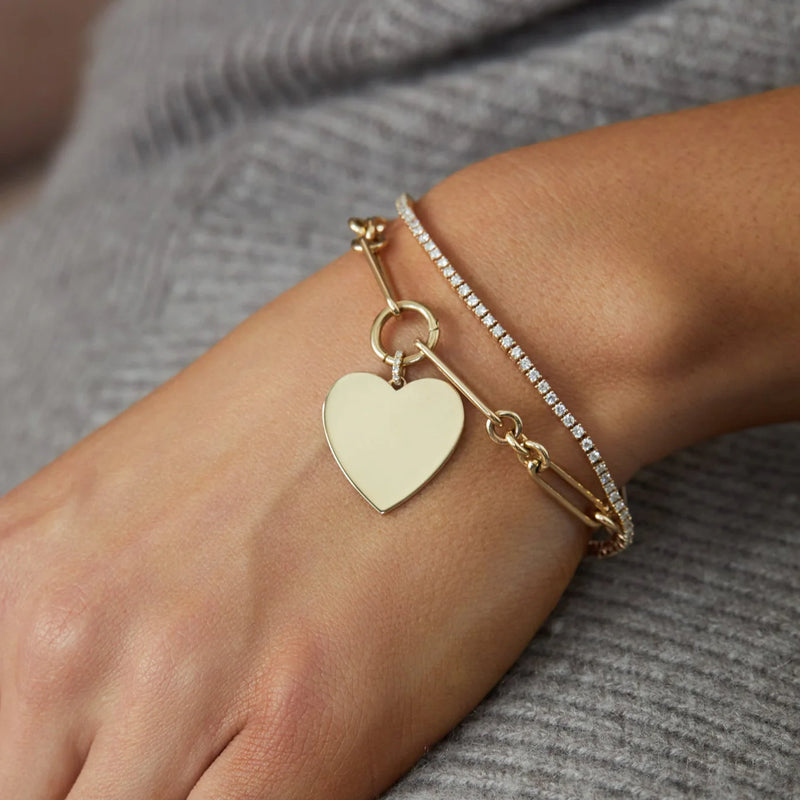 A close-up features a persons wrist wearing an ef collection Gold Jumbo Heart Charm Bracelet alongside a silver bracelet with small crystals, both elegantly resting on gray textured fabric.