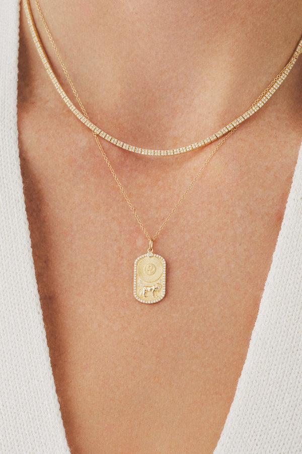 A close-up of a person wearing two gold necklaces—one thin chain and one ef collection Diamond Zodiac Charm—over a white textured V-neck top.