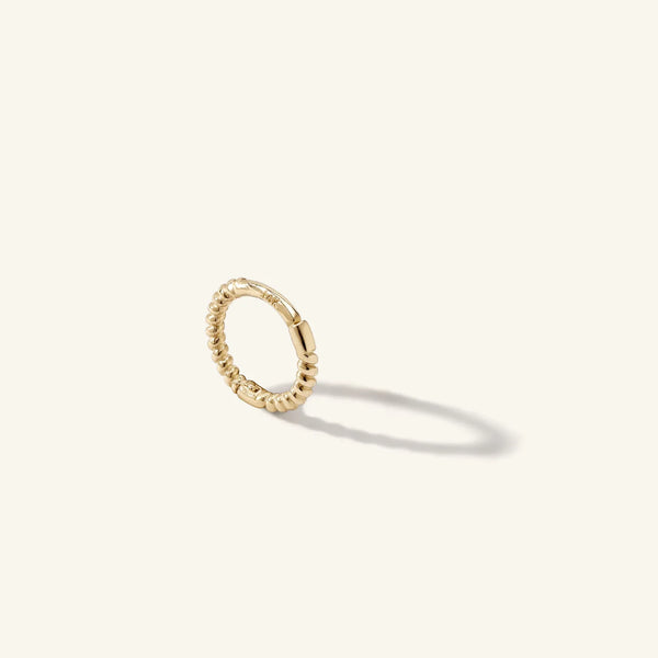 A gold Earring Charm Necklace Connector by zahava, featuring bead-like segments, stands upright against a plain, light backdrop. It casts a soft shadow to the right, highlighting its textured details.