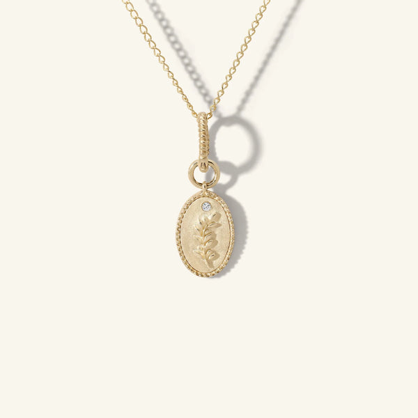 A Zahava Earring Charm Necklace Connector features a delicate chain with an oval pendant engraved in a floral design. Accented by a pavé diamond, the pendant shines brilliantly against a plain, light background.