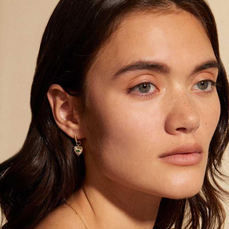 A woman with long brown hair gazes to the side, highlighting a small gold heart-shaped earring and wearing the zahava Mini Emerald Sunbeam Heart Charm necklace. The background is a neutral beige color.