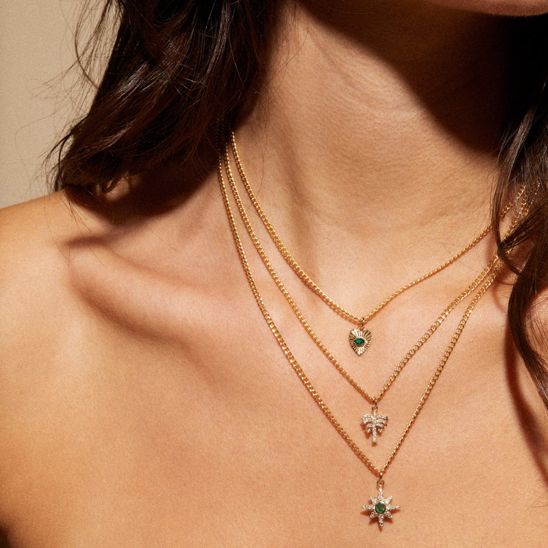 Wearing a Zahava Mini Emerald Sunbeam Heart Charm, a woman adorns herself with three-layered 10k yellow gold necklaces featuring pendants of a heart, cluster, and starburst. Each pendant sparkles with small green gemstones. Her style pairs gracefully with an off-the-shoulder top.