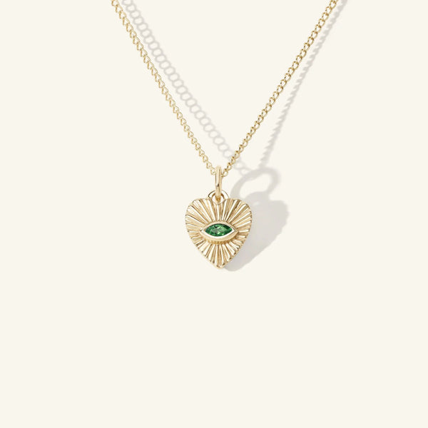 The Mini Emerald Sunbeam Heart Charm by zahava features a 10k yellow gold heart pendant with a central green eye and a sunburst pattern. The charms delicate chain beautifully rests against a plain, light background, capturing attention with its elegant design.