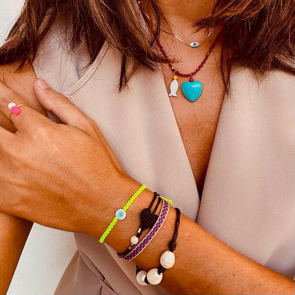 A woman with tan skin wears the Oiya Alora Turquoise Heart Necklace layered with a blue heart pendant, plus colorful beaded, evil eye, and red lapis stone bracelets, styled with a beige sleeveless top.