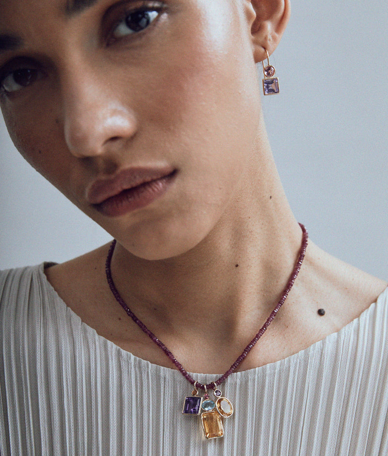 Wearing a pleated, light top and Lizzie Fortunatos Tiny Beaded 14k Gold Necklace in Ruby with matching gemstone earrings, a person gazes softly at the camera.