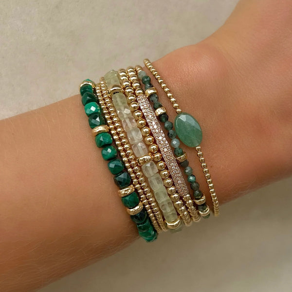 A wrist showcases multiple handmade bracelets from Los Angeles, including the May Emerald and Rondelle Bracelet by Karen Lazar, featuring green, white, and gold beads in diverse textures and designs. Some bracelets include small round beads, while others are embellished with 14k yellow gold accents or emerald gemstones for a layered and stylish look.