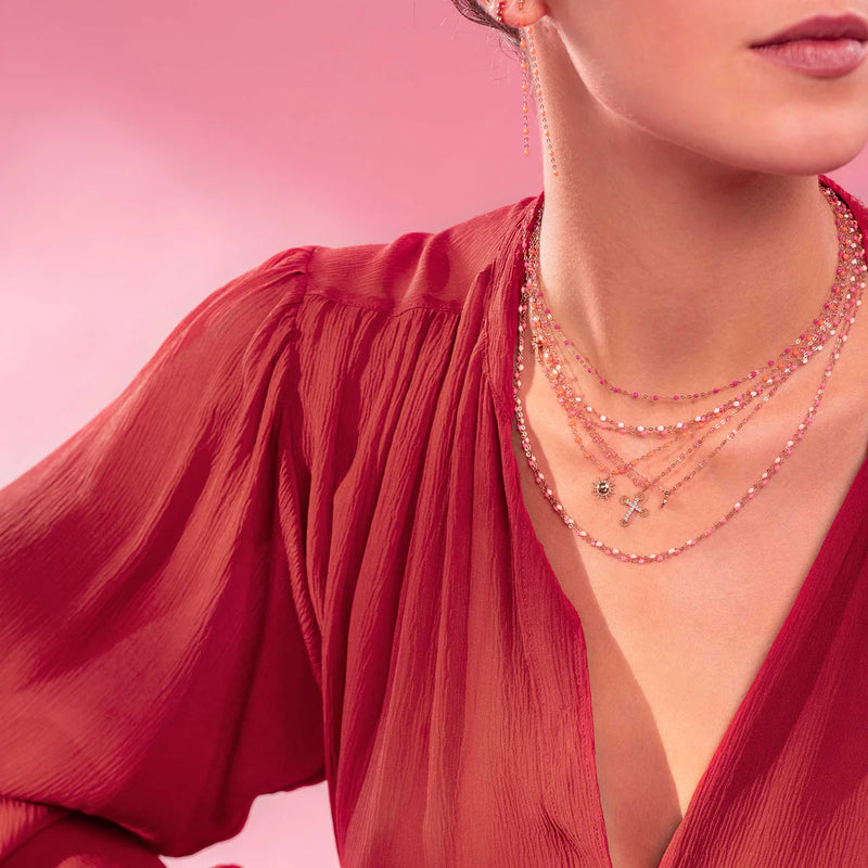 A woman in a deep red, pleated blouse and layered gold necklaces—including the Gigi Clozeau Sun Pendant—poses against a pink background, revealing only her lower face, neck, and upper torso.
