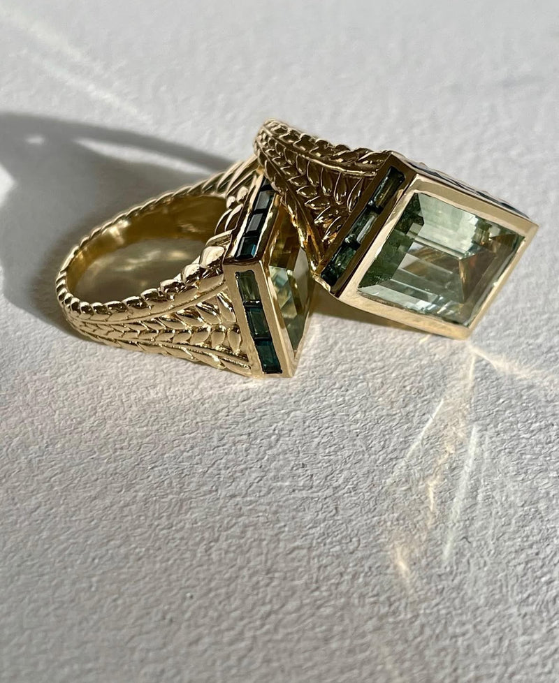 Two Pascale Monvoisin Poppy Ring Green Amethyst signet rings in yellow gold, each with a textured band and large rectangular green amethyst gemstone, rest on a white surface casting soft shadows and reflections.