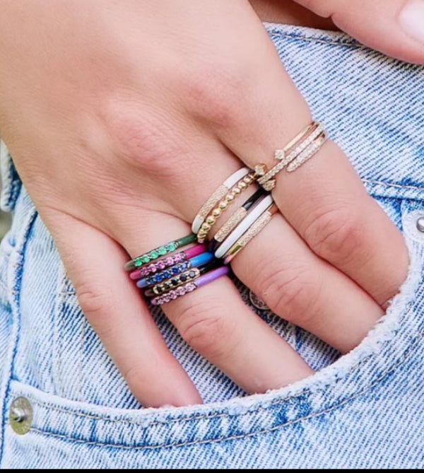 A hand adorned with various colorful rings, including a Black Enamel And Spinel Band Ring by Rachel Reid and 14K yellow gold and gemstone bands, rests on the pocket of light blue denim jeans.