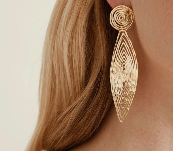 Close-up of a womans ear adorned with gas bijoux Longwave earrings large size, featuring gold-plated, Mediterranean-inspired spiral and teardrop shapes. Her light blonde hair is tucked back, showcasing the handcrafted jewelry against a neutral background.