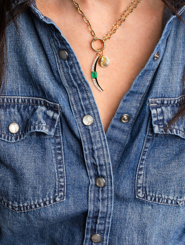 Someone in a denim button-up shirt wears Monarch Markets Diamond Enamel Moon, a gold chain necklace featuring a hoop pendant, seashell charm, and an emerald-accented crescent moon with green detailing.