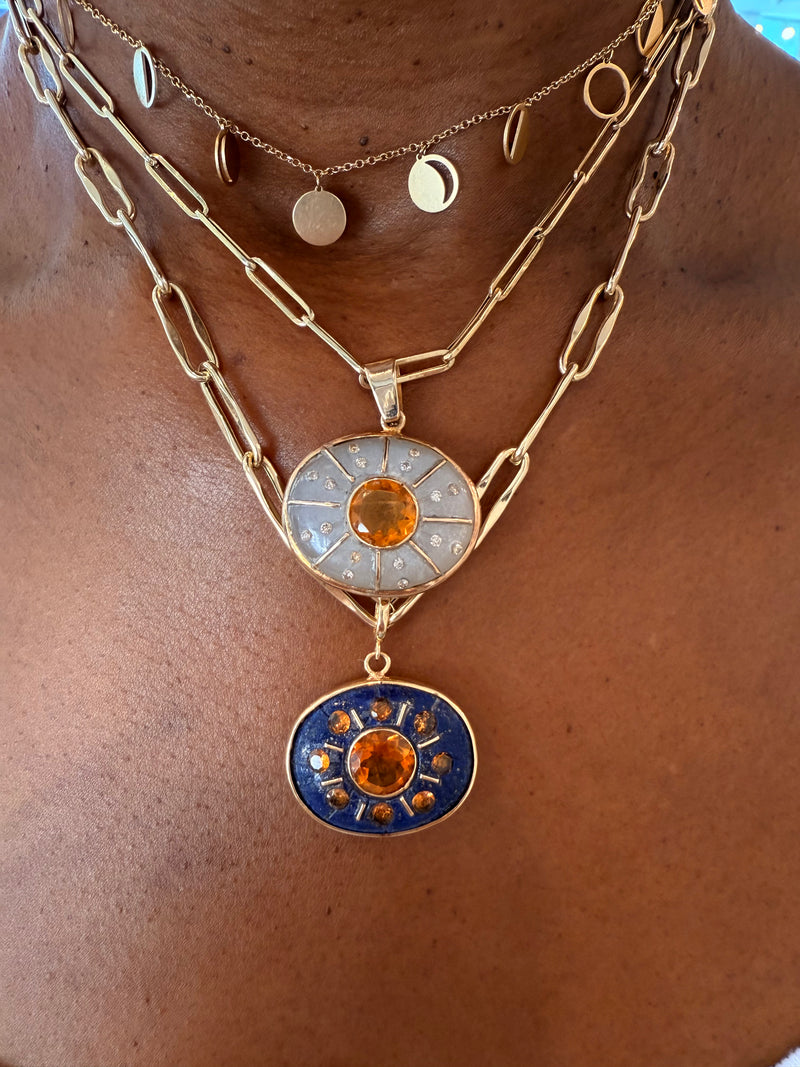A close-up of a person wearing layered gold necklaces, including Jen Proudman's SOL Pendant featuring moonstone, citrine, and diamonds, along with other pendants in gold.
