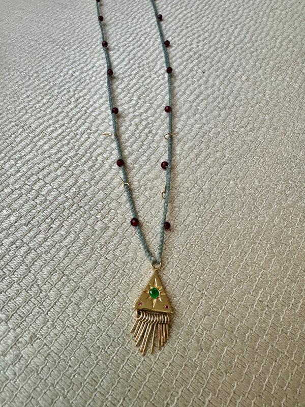 The Nanyehi Charm Necklace by Scosha showcases a solid gold build with a thin blue cord accented by small red beads. A gold triangular pendant featuring an emerald garnet gemstone and fringed design hangs at the center, elegantly displayed on a neutral textured surface.