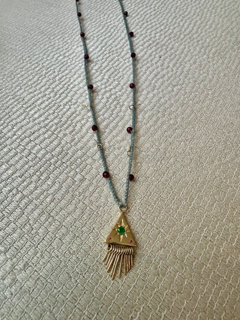 The Nanyehi Charm Necklace by Scosha showcases a solid gold build with a thin blue cord accented by small red beads. A gold triangular pendant featuring an emerald garnet gemstone and fringed design hangs at the center, elegantly displayed on a neutral textured surface.