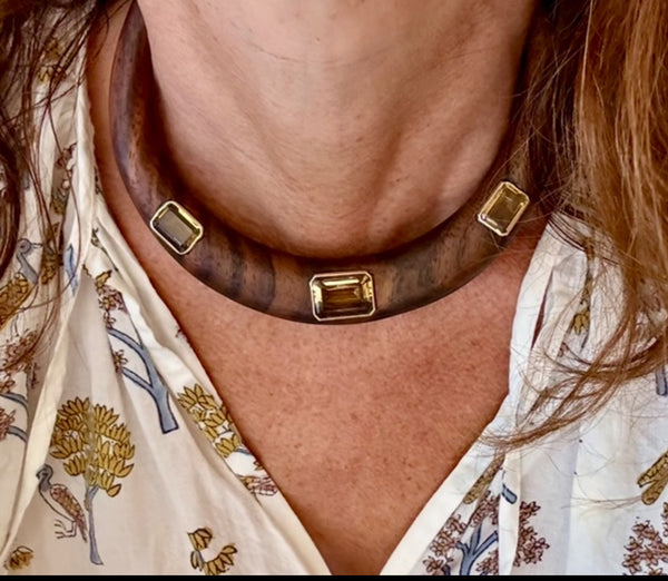 A person wears the Jen Proudman Wood and Citrine Collar, a thick dark wood necklace with 14k gold accents, paired with a white patterned blouse decorated with trees and birds.