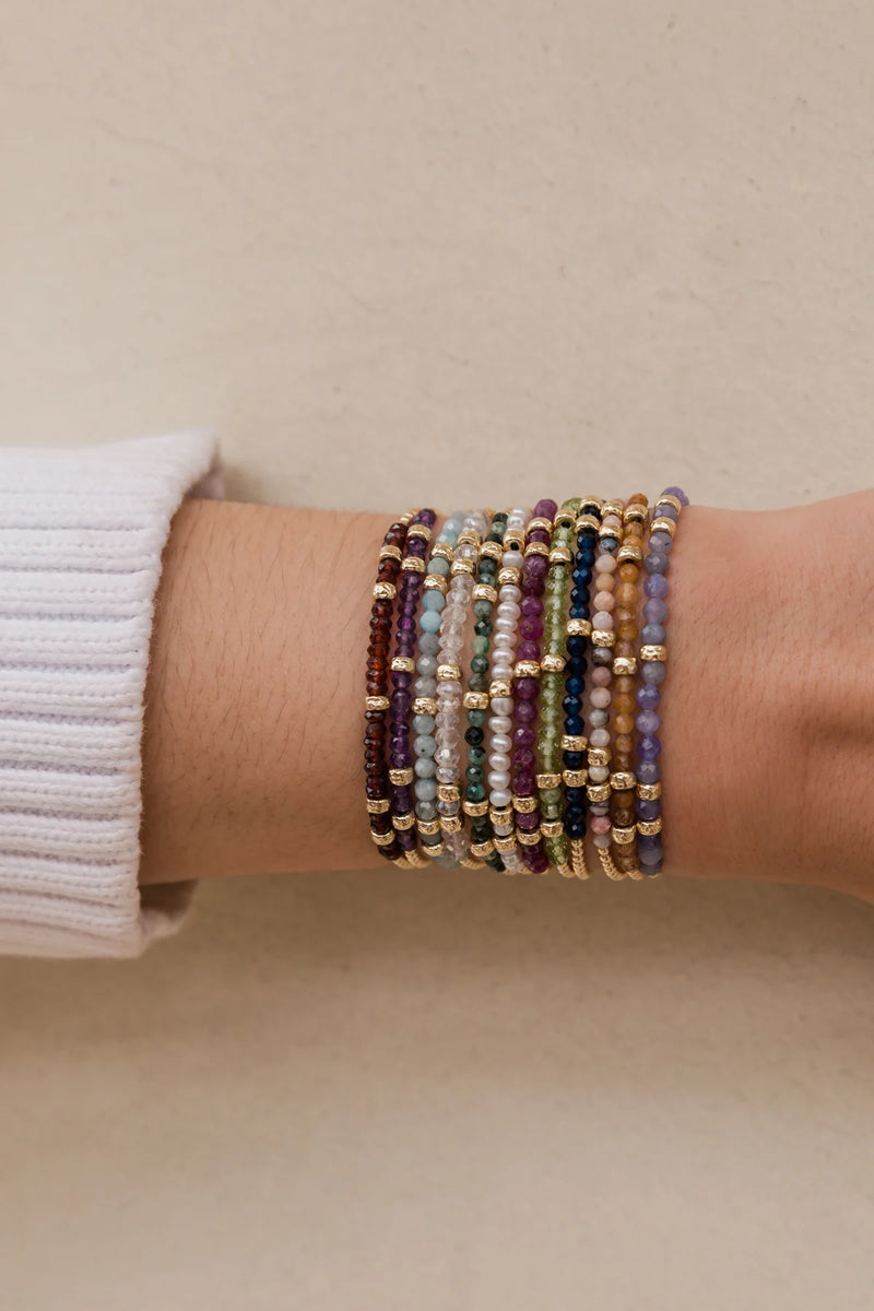 A person's wrist adorned with the Karen Lazar May Emerald and Rondelle Bracelet, along with other vibrant beaded accessories, showcases shades of purple, emerald green, and gold with metallic accents. Crafted with love in Los Angeles, they pair these bracelets elegantly with a white long-sleeve top.