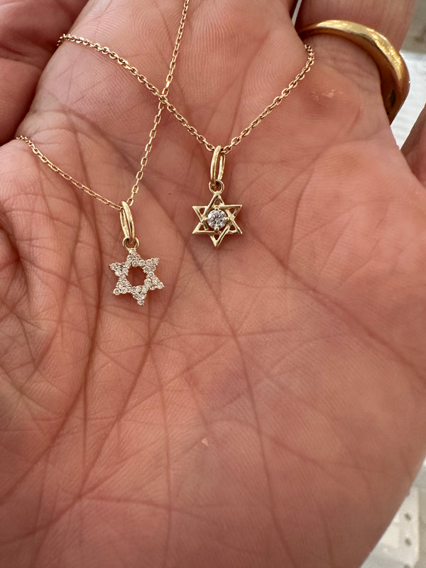 A close-up of a hand holding two Rachel Reid Mini Jewish Star Charms in 14k gold—one with diamond accents, the other with a central gemstone; a gold ring is worn on one finger.