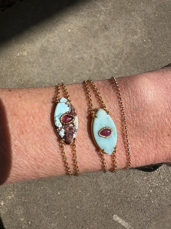 A wrist showcases two AYIN Bracelet Larimar pieces by Bohemian Rhapsodie Paris, each handcrafted with an oval turquoise stone and a central reddish-purple gem, gleaming in the sunlight on a textured surface.
