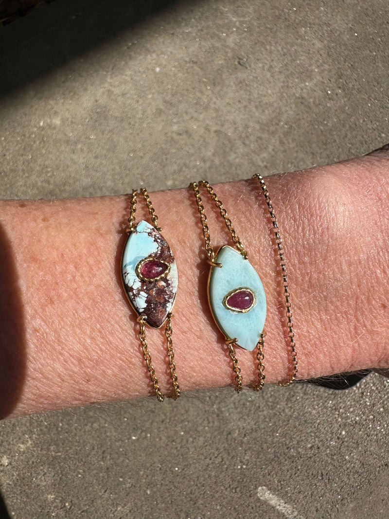 A wrist showcases two AYIN Bracelet Larimar pieces by Bohemian Rhapsodie Paris, each handcrafted with an oval turquoise stone and a central reddish-purple gem, gleaming in the sunlight on a textured surface.