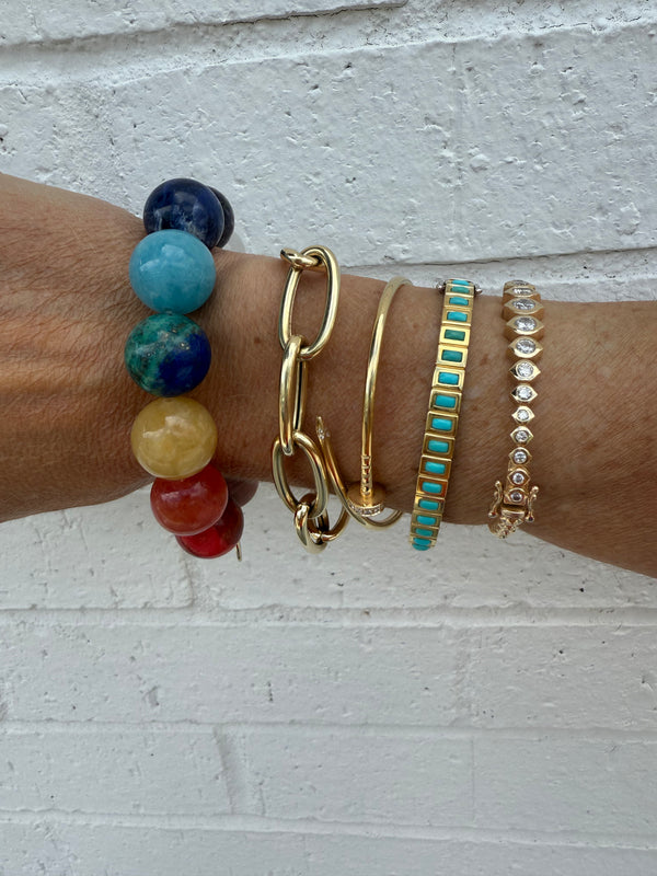 A wrist styled with five bracelets, including Monarch Market’s Rainbow Gemstone Bracelet, against a white brick wall. The stack features colorful beads, a thick gold chain, a thin bangle, turquoise-accented gold, and delicate gold jewelry.