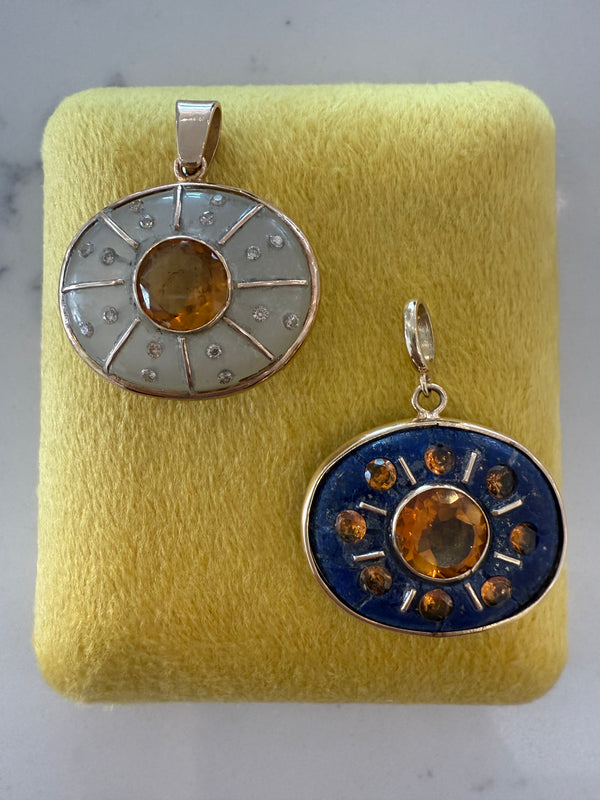 Two circular SOL Pendants by Jen Proudman—one with moonstone, the other with citrine and diamonds—are displayed on yellow fabric.