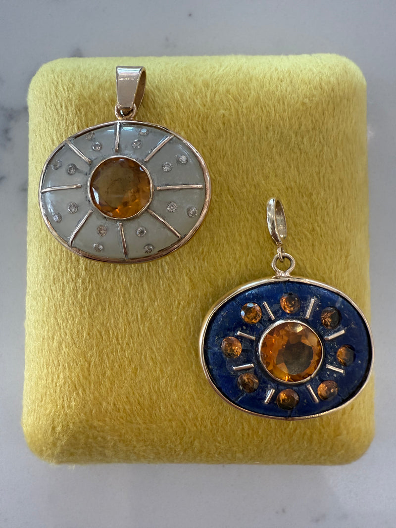 Two circular SOL Pendants by Jen Proudman—one with moonstone, the other with citrine and diamonds—are displayed on yellow fabric.