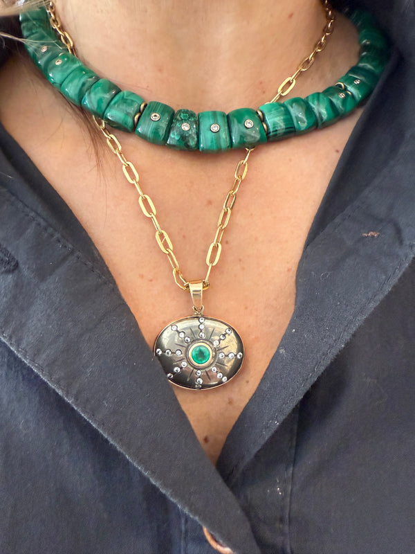 A person wears the Jen Proudman Sol Pendant—Pyrite, Emerald, and Diamonds—featuring a gold chain and round 14k gold pendant with emerald, over a dark collared shirt.