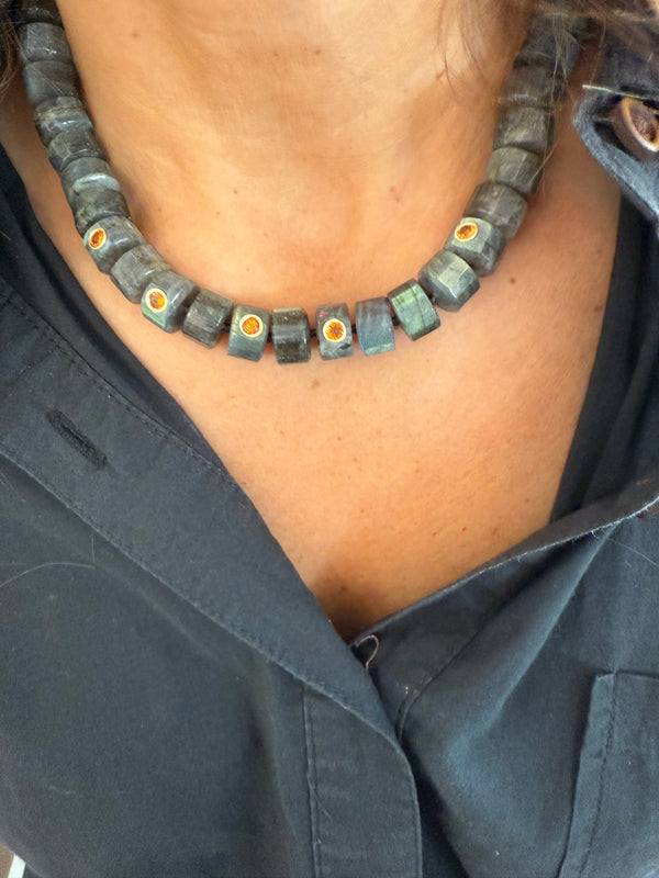 A close-up shows a person in a dark blue collared shirt wearing the Jen Proudman Grand Labradorite Necklace, a chunky handmade piece with dark beads and gold/yellow accents.