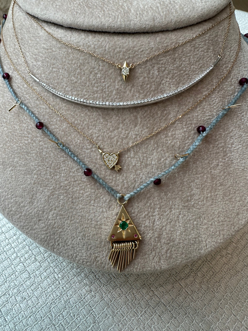 A display showcases five delicate Scosha Nanyehi Charm Necklaces: a solid gold sparkling bar, a star pendant, a heart with wings, a blue string with red beads, and a triangle pendant with an emerald garnet and gold tassels.