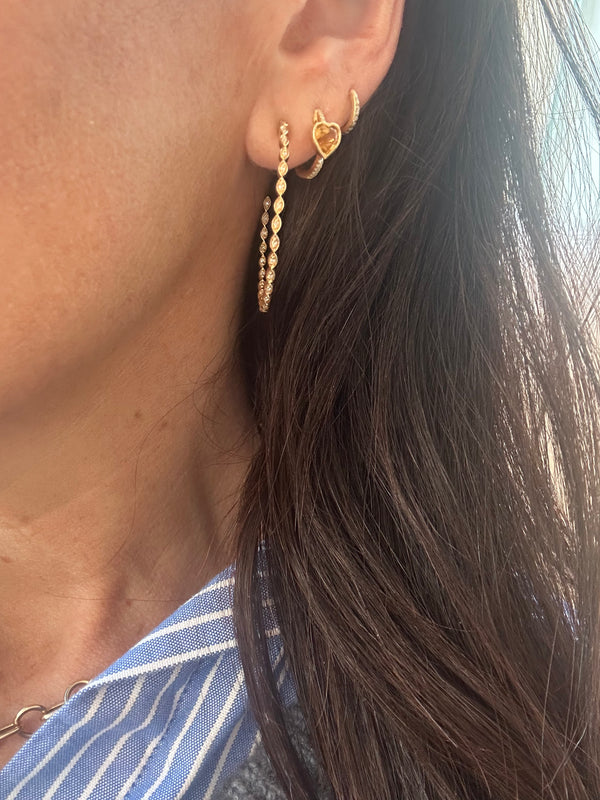 Close-up of a person wearing Rachel Reids Gold and Diamond Marquise Hoops along with two other 14k yellow gold earrings: a smaller hoop with heart pendant and a simple twist design. The person has long dark hair and wears a blue-and-white striped shirt.