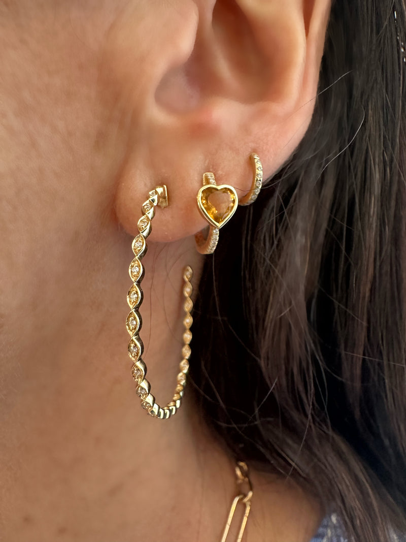 A close-up of a womans ear features Rachel Reids Gold and Diamond Marquise Hoops: one large 14k yellow gold hoop with an intricate design and a smaller marquise hoop with a heart shape center. Her dark hair, partially visible, enhances the elegance.