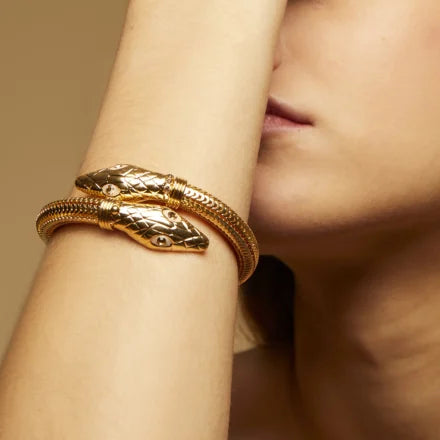 A woman with light skin wears the Cobra Bracelet by gas bijoux, a gold-plated statement piece featuring two detailed snake heads facing each other, wrapped around her wrist. Her face is partially visible, and her arm is raised.