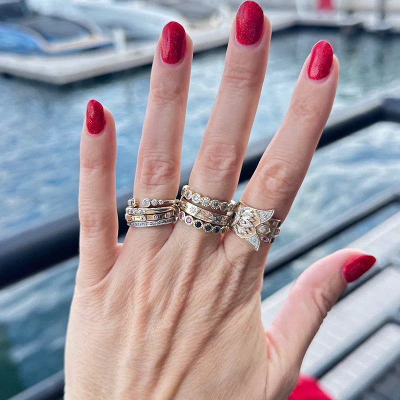 A hand with red nail polish displays an array of gold and diamond rings, featuring a Bejeweled Sideways Hamsa Ring by Three Stories Jewelry, set against a scenic waterfront with boats.