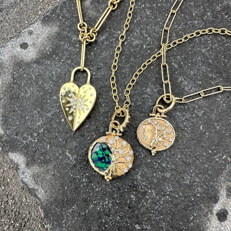 Three gold necklaces rest on a stone surface. The first, featuring a heart-shaped locket with floral designs, elegantly hangs from Three Stories Jewelrys Classic Medium Textured Oval Chain. The other two have circular pendants: one with a colorful stone and the other in 14k yellow gold with intricate tree motifs.