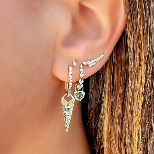 Close-up of an ear with multiple earrings from Three Stories Jewelry. A large geometric earring featuring colorful gems and a smaller green gem dangles below, complemented by a 14k yellow gold stud and a Long Pave Ear Crawler with intricate wing design and pave diamonds. Hair partially visible.