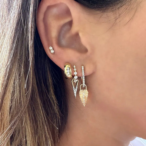 Close-up of an ear showcasing multiple piercings: a small stud, a 14k yellow gold hoop, a triangular dangle, one with diamond-adorned leaf design, and Three Stories Jewelrys Classic Open Dagger Charm.