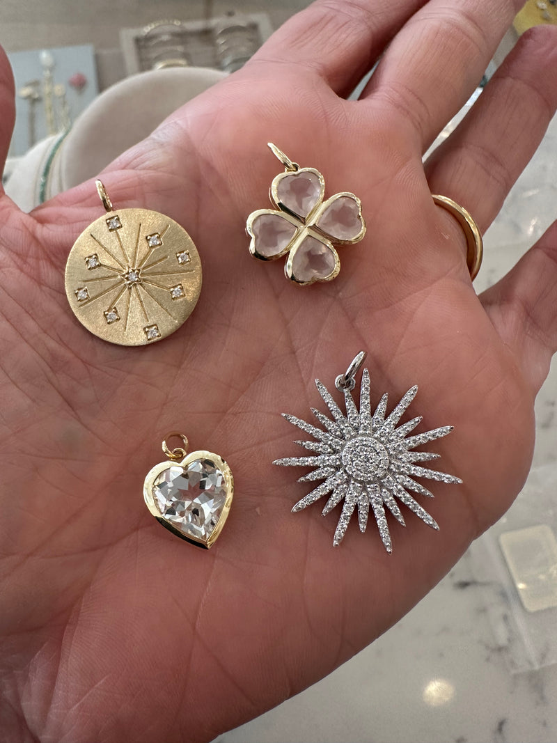 A hand holds four pendants: a 14k yellow gold circle with diamond accents, a gold flower with pale pink petals, Rachel Reids Gemstone Heart Charm in a gold frame, and a silver sunburst adorned with White Topaz.