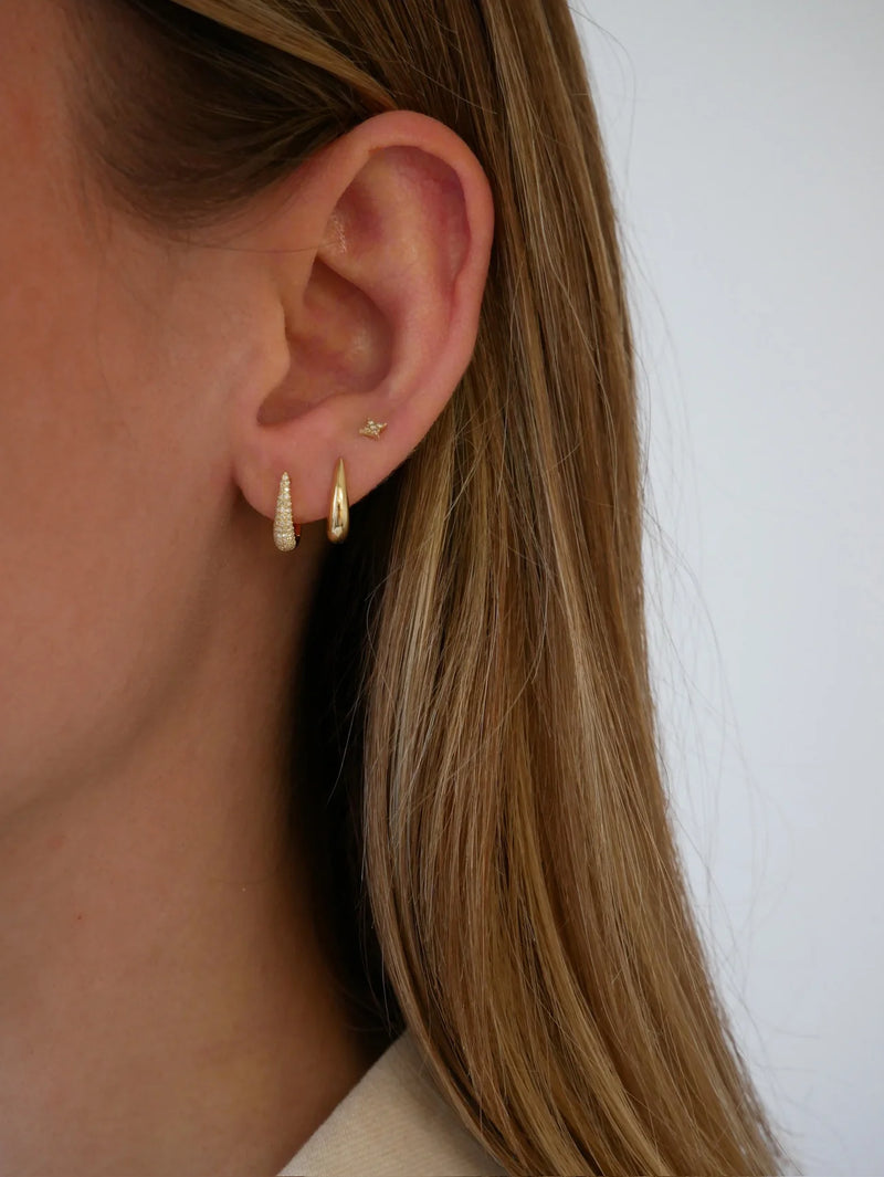 Close-up of a womans ear wearing ef collection Gold Mini Maeve Huggie Earrings in 14k yellow gold, paired with a star stud and a plain hoop. Her straight, light brown hair frames this elegant earring arrangement.