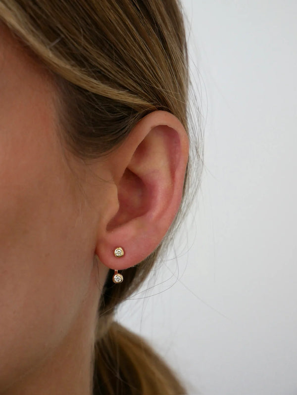 Close-up of a womans ear wearing ef collections Diamond Pillow Illusion Earrings—minimalist, round studs with clear bezel-set diamonds—adding a contemporary touch against a plain, light background.