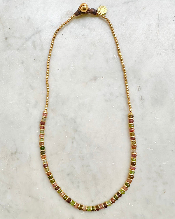 The CHOKER ELLIE by Bohemian Rhapsodie Paris is elegantly displayed on a marble surface, featuring small gold beads seamlessly intertwined with larger multicolored tourmaline beads in shades of green, red, and brown, all secured with a gold clasp. It's perfect for meditation or adding an earthy touch to any ensemble.