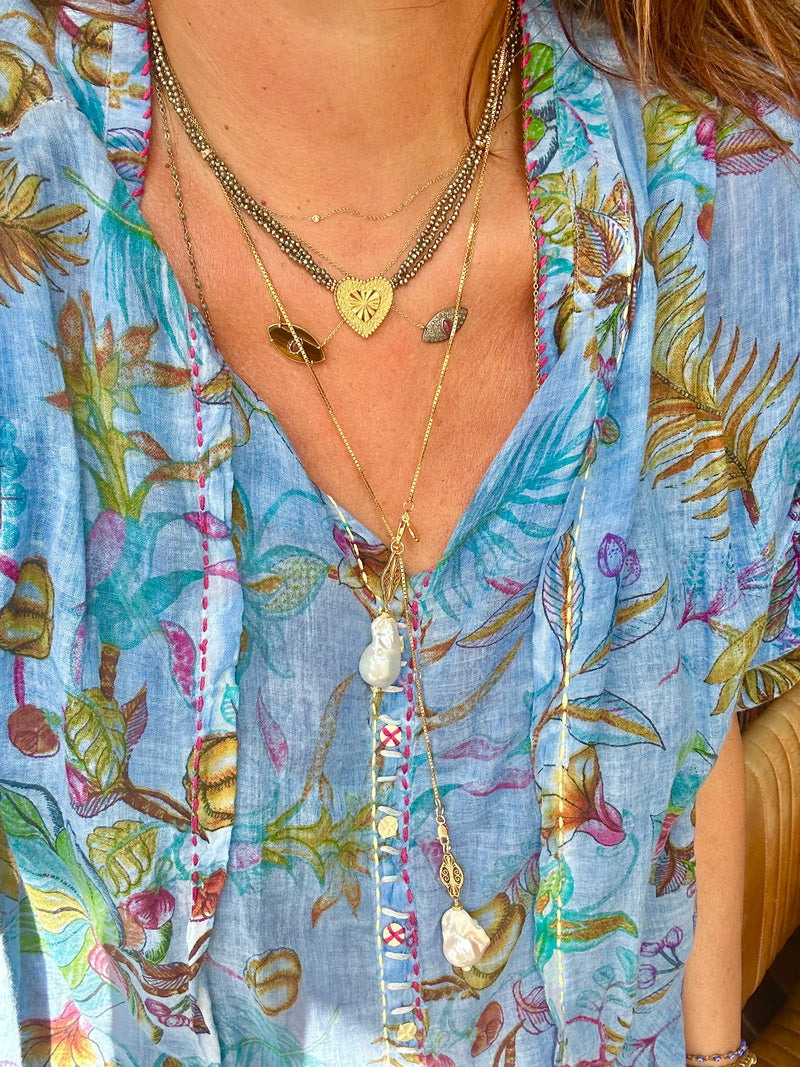 A person is dressed in a light blue floral top with colorful embroidery, accessorized with multiple layered necklaces. Among the jewelry collection, they showcase the JOHN ROSS choker from Bohemian Rhapsodie Paris, adorned with a multicolored tourmaline pendant. This piece beautifully complements gold-plated heart details and various gemstones.