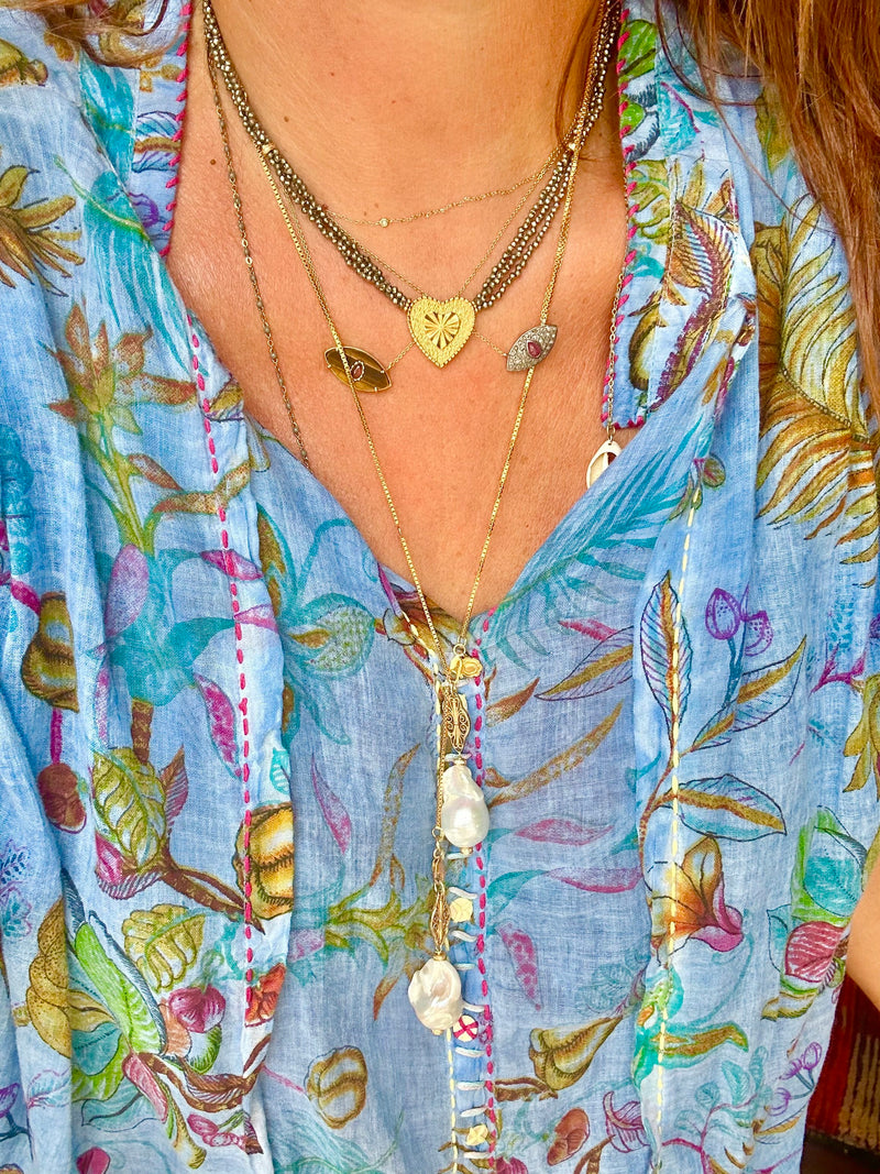 A person wearing a JOHN ROSS blue floral-patterned shirt from Bohemian Rhapsodie Paris, featuring vibrant leaf and flower designs. Around the neck are several layered necklaces, including the ELLIE Choker adorned with multicolored tourmaline and pearls. Gold-plated charms and heart shapes add an elegant touch.