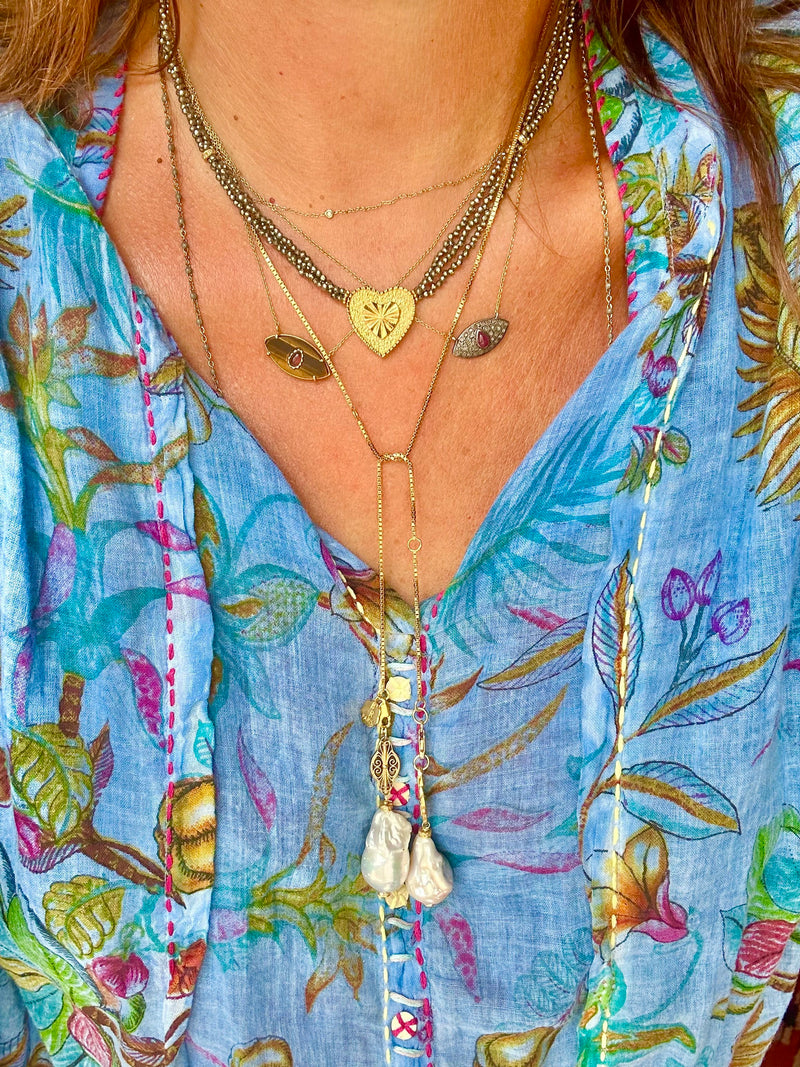 A person wears layered gold-plated necklaces by Bohemian Rhapsodie Paris, including heart and eye pendants adorned with multicolored tourmaline, along with an ELLIE Choker. They are dressed in a vibrant blue floral-patterned top showcasing colorful stitching.