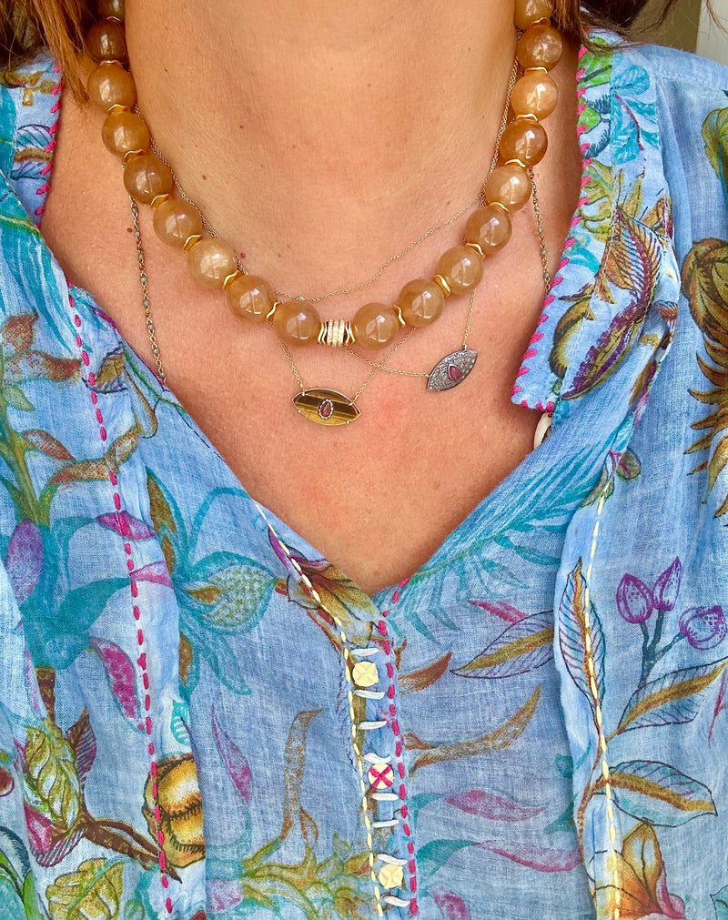 A person wearing a colorful floral-patterned blouse with red stitching, adorned with layered necklaces featuring a beaded design and pendant charms, highlights one piece from Bohemian Rhapsodie Paris with a gold-plated chain and another showcasing the jade SLY Camel charm.