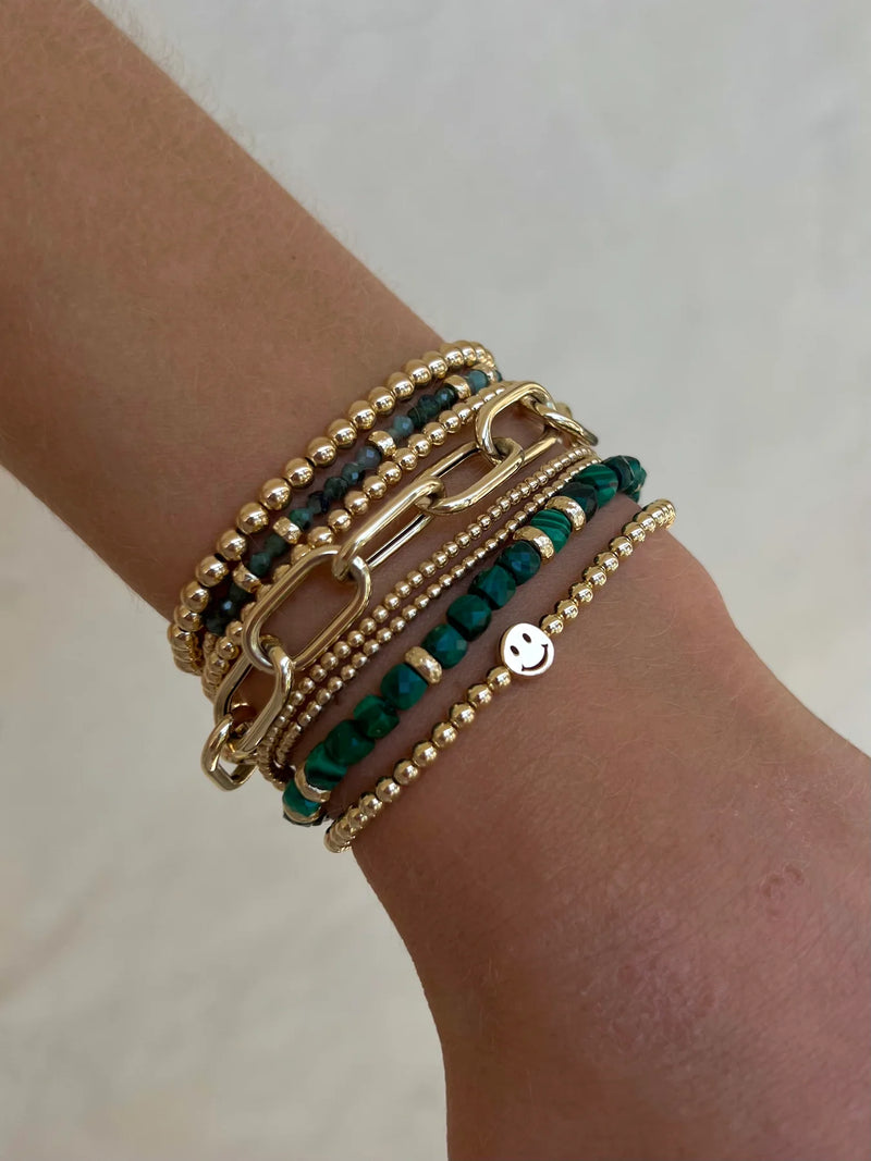 A wrist featuring multiple bracelets, such as a May Emerald and Rondelle Bracelet by Karen Lazar, which includes gold bead strands, a 14k yellow gold chain link bracelet, and vibrant green emerald gemstone beads. Additionally, there is a small round charm with a smiling face. Each piece is meticulously handmade in Los Angeles.