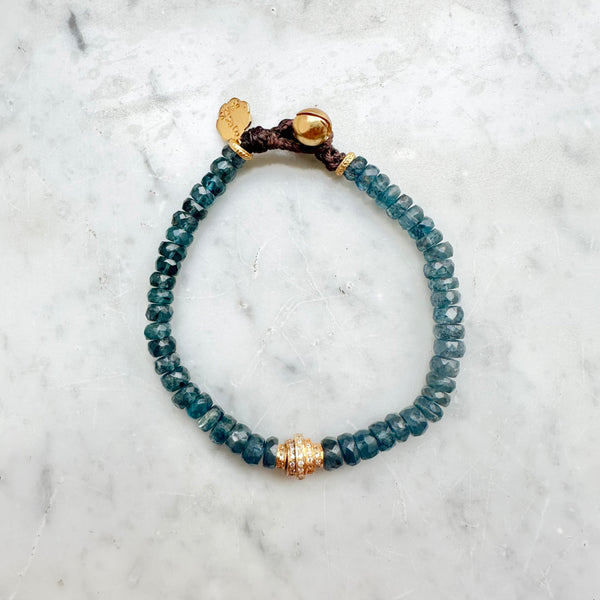 The CLAYTON Bracelet by Bohemian Rhapsodie Paris features translucent teal beads with a central labradorite-accented bead, secured by a brown cord adorned with a gold bead and a small tag, elegantly displayed on a light marble surface.