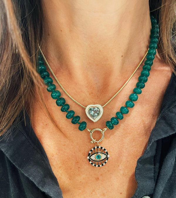 A person wears two necklaces: the Protection Charm by Kate Collins Jewelry, featuring a green beaded chain with an emerald eye charm, and another with a thin 14 Karat Yellow Gold chain and heart-shaped pendant. This ensemble is perfectly complemented by their dark shirt.