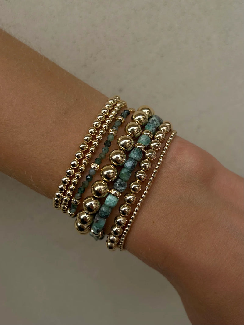 A wrist is adorned with multiple Karen Lazar bracelets, including the May Emerald and Rondelle Bracelet featuring 14k yellow gold, small gold beads, and vibrant turquoise accents. Stacked together against a light background, each piece is meticulously handmade in Los Angeles, creating an effortless blend of luxury and artistry.